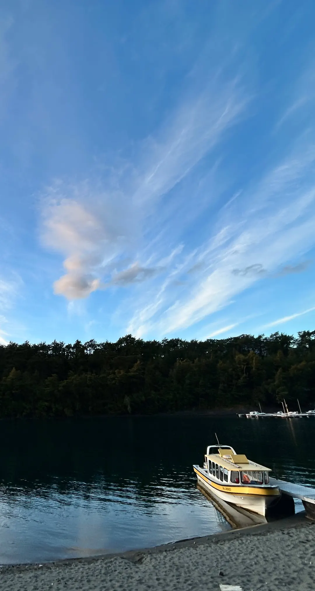 La lancha amarilla amarrada en la orilla del lago, con el bosque y el volcán Osorno nevado detrás, bajo un cielo de nubes largas.