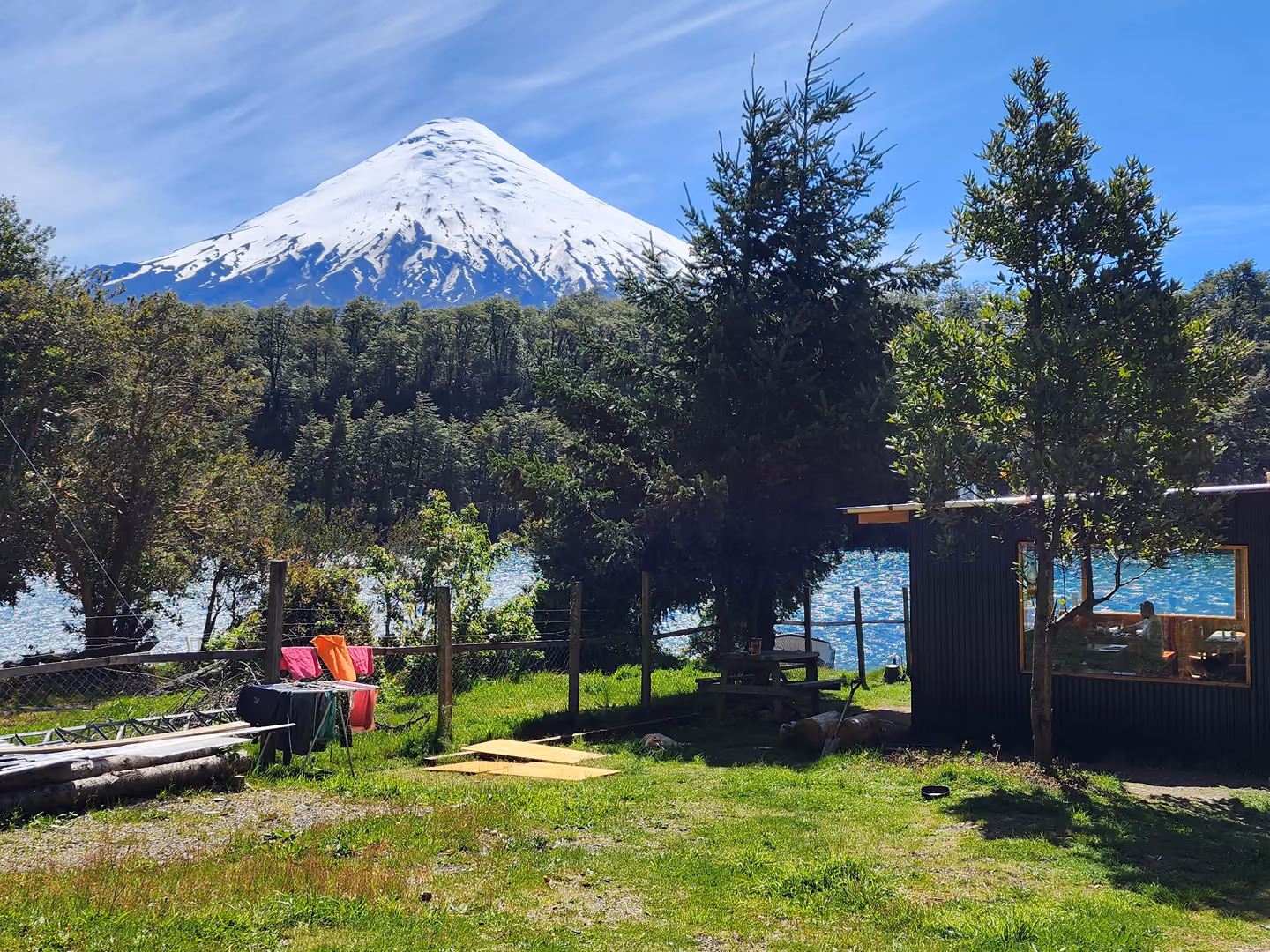 El volcán Osorno nevado asoma sobre los árboles; abajo, el pasto y el comedor de madera oscura con techo de zinc.