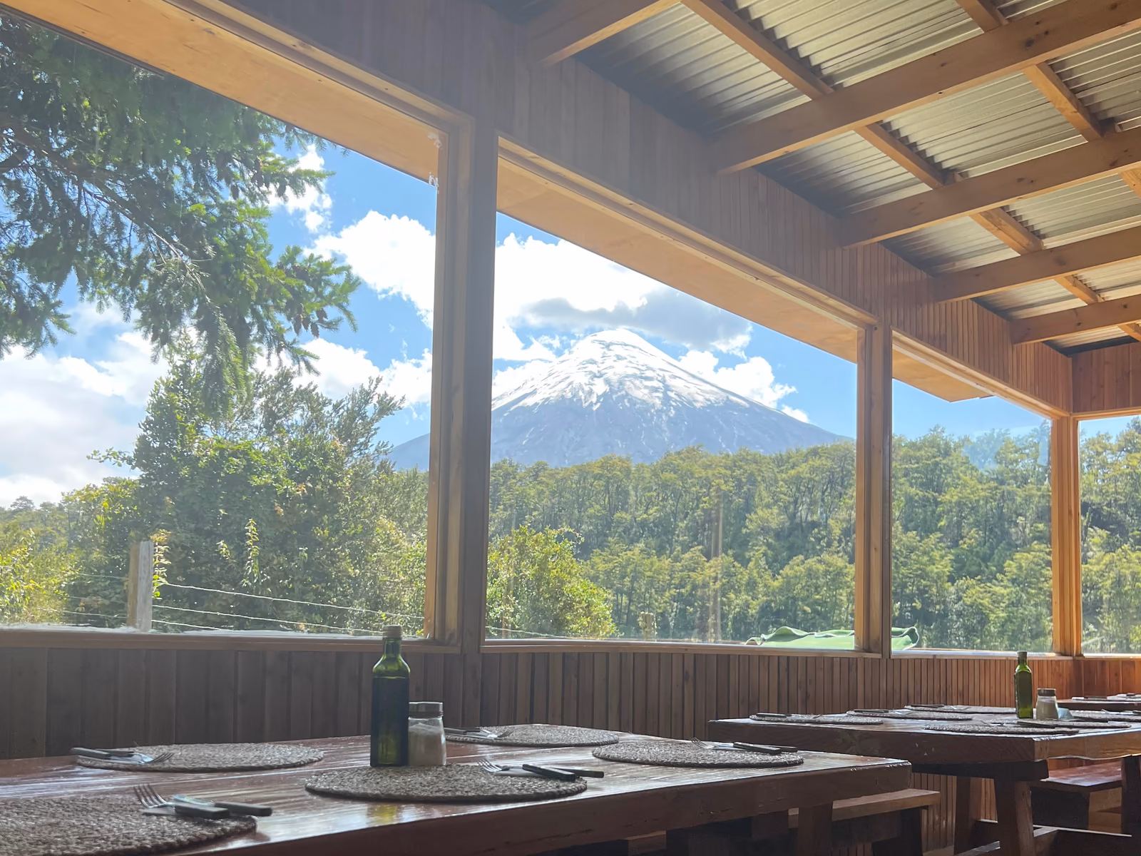 El comedor por dentro: mesas de madera con individuales tejidos y, por el ventanal, el volcán Osorno sobre el bosque.