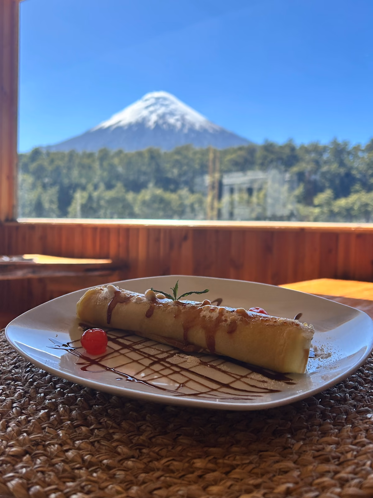 Postre en un plato blanco sobre un individual tejido; detrás, por el ventanal, el volcán Osorno sobre el bosque.